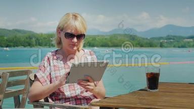 假期的技术。 一位拿着平板电脑的女士坐在湖和山背景的咖啡馆里的桌子旁